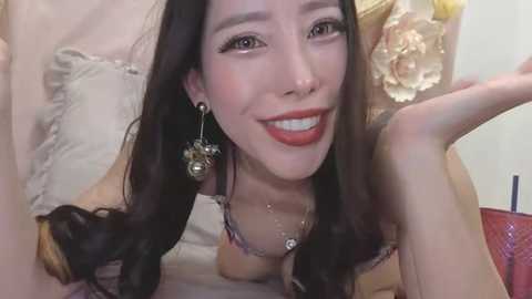 Media: A video of an Asian woman with long black hair, wearing red lipstick, earrings, and a necklace, lying on a bed with a beige pillow, smiling.