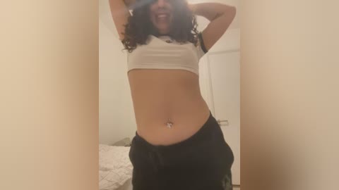 Media: A video of a light-skinned woman with curly hair, wearing a cropped white top and black pants, taking a mirror selfie in a dimly lit room with beige walls.