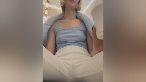 Media: Video of a blonde woman with a slim build, wearing a light blue tank top and white pants, seated in an airplane seat, her legs spread wide, capturing a provocative and suggestive pose.