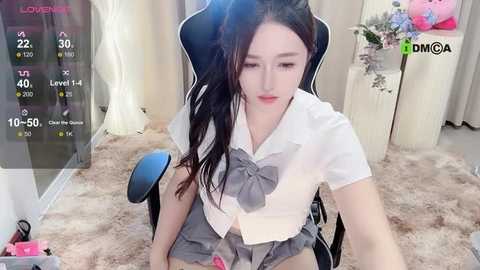 Media: Video of a young Asian woman with long black hair, wearing a white blouse and grey skirt, sitting on a black chair in a cozy, dimly-lit room.