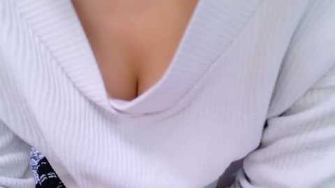 Media: Close-up video of a woman's chest in a white, ribbed, V-neck sweater, revealing cleavage and a hint of a black bra. Her skin is light, and the focus is on the fabric texture and neckline.