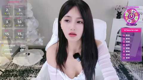 Media: Video of an East Asian woman with long black hair, fair skin, and a slender physique, wearing a white off-shoulder top, sitting on a white chair in a minimalist room with a black and white patterned rug.