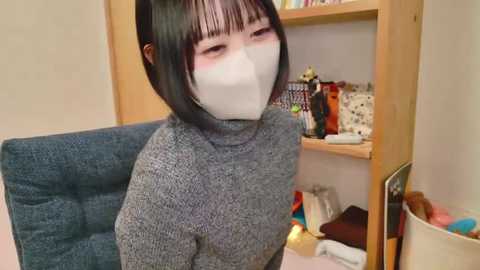 Media: Video of an Asian woman with short black hair, wearing a grey turtleneck sweater, white mask, and gag, sitting on a blue chair in a cluttered room with a wooden shelf, stuffed toys, and a wicker basket.