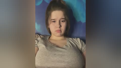 Media: Video of a young Caucasian girl with light skin and a ponytail, wearing a gray T-shirt. She appears sad, with a neutral background of blue and purple tones.