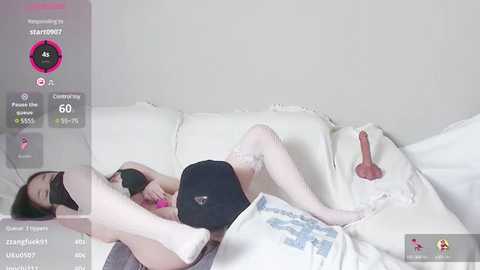 Media: Video of a woman lying on a white couch, wearing a black bra, black shorts, and white thigh-high stockings. The background is a plain, light-colored wall.