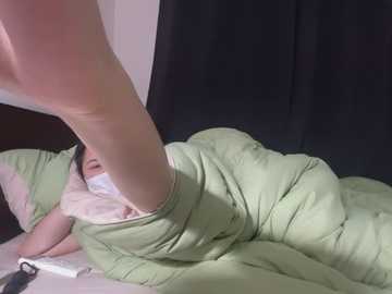 Media: A video of a person lying in bed, partially covered by a light green blanket, with a visible leg extended, set against dark curtains and a white wall.