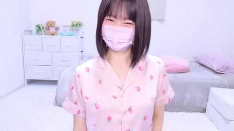 Video of a young East Asian woman with short black hair, wearing a light pink button-up shirt adorned with small strawberry patterns, a pink face mask, and sitting in a minimalist, pastel-toned bedroom.