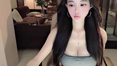 Media: Video of an East Asian woman with long black hair, wearing a strapless gray top, standing indoors next to a dark sofa and glass doors.