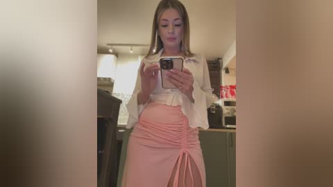 Media: Video of a pregnant woman with light skin, straight blonde hair, wearing a white blouse and pink, ruffled skirt, taking a selfie in a modern kitchen.