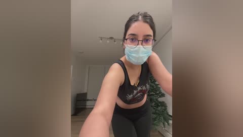 Media: A video of a young woman with light skin, brown hair, and glasses, wearing a black sports bra and leggings, taking a selfie in a modern, minimalist living room.