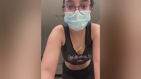 Media: Video of a young woman with light skin, wearing glasses, a blue surgical mask, and a black sports bra with a graphic design, leaning forward. Background is a dimly lit room with neutral walls.