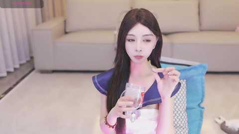 Media: Video of a slender Asian woman with long black hair, dressed in a sailor schoolgirl outfit, drinking wine indoors. Background features a beige couch, white curtains, and a blue cushion.