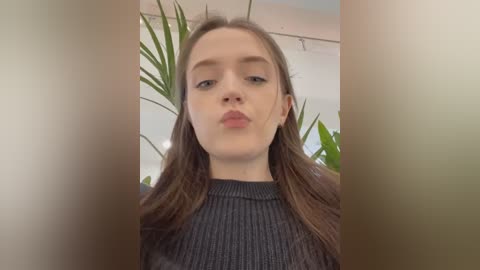 A video of a young Caucasian woman with fair skin and long brown hair, wearing a dark gray ribbed sweater, blowing a kiss into the camera. Background features green potted plants and white walls.