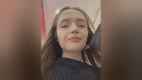 Media: Video of a young woman with fair skin, long light brown hair, and closed eyes, wearing a black sweater, taken from a low angle. Background is blurry with a hint of red and white.