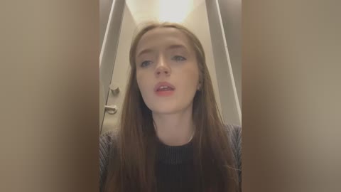 Media: A video of a young Caucasian woman with long, straight brown hair, fair skin, and light makeup, wearing a black top, standing in a narrow hallway with beige walls and a white door.