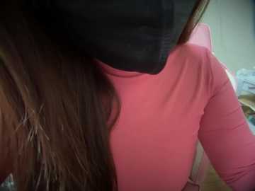 Media: A close-up video of a person wearing a pink long-sleeve shirt and a black mask, with long, dark brown hair visible on the left side. The background is blurred, showing a wall and indistinct objects.