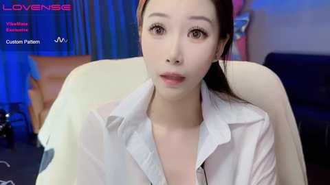 Media: Video of a young Asian woman with fair skin, wearing a white button-down shirt, sitting on a beige couch. The background features blue curtains and a dark couch.