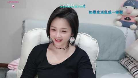 Media: Video of a young Asian woman with short black hair, wearing a black top, seated on a light blue sofa, smiling. Background includes stuffed toys and text in Chinese.