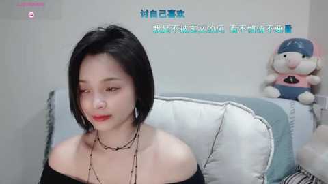 Media: Video of an Asian woman with short black hair, wearing a black off-shoulder top and multiple necklaces, seated on a white couch with a plush toy in the background.