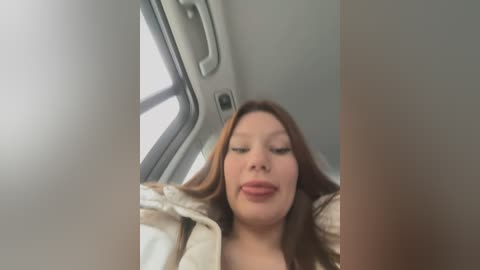 Media: A video of a woman with light skin, brown hair, and closed eyes, wearing a beige jacket, seated inside a car with a gray interior.