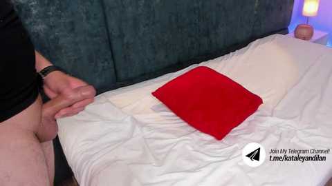 Media: Video of a man with a circumcised penis standing at the edge of a bed with a white sheet and a red pillow, wearing black shorts. Background includes a teal headboard and a bedside lamp.