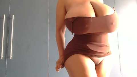 Video of a curvaceous woman with large breasts, wearing a strapless, form-fitting, brown dress, standing in front of modern grey cabinets with metallic handles.