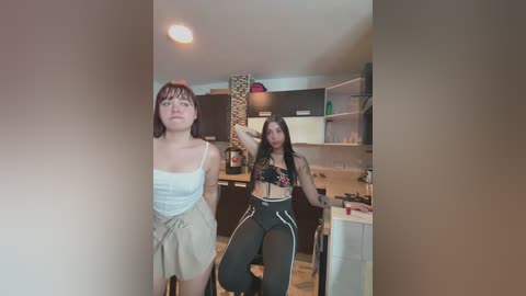 Media: Video of two women in a kitchen: one with short hair in a white tank top, the other with long hair in a black sports bra and high-waisted pants.