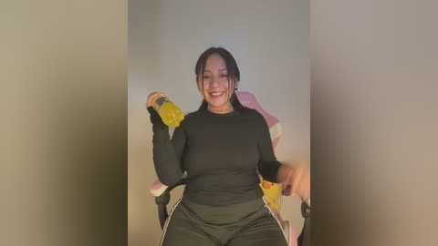 Media: Video of a smiling, young Asian woman with black hair, wearing a black long-sleeve top and black pants, sitting in a pink gaming chair, holding two yellow energy drinks.