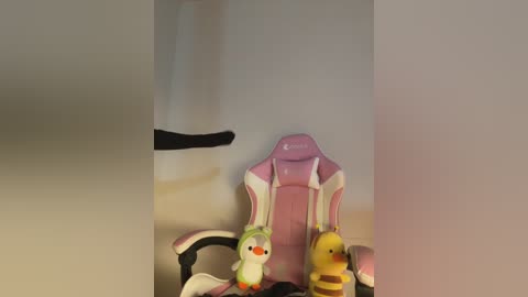 Media: A video of a pink gaming chair with two small, decorative toy birds\u2014one green and one yellow\u2014sitting on the armrests. The background is blurred and off-white.