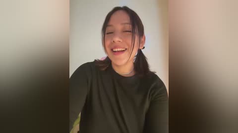 Media: Video of an Asian woman with medium-length black hair, smiling joyfully. She wears a black top, standing in a blurred, beige hallway.