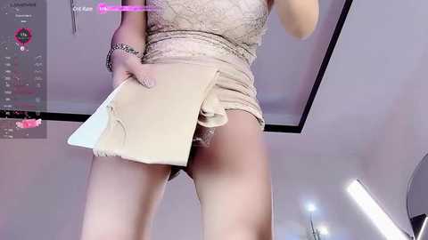 Media: Video of a woman in a short, beige lace dress, exposing her buttocks, holding a beige clutch bag, taken from a low angle.