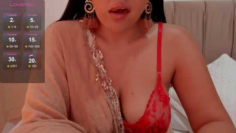 Media: Video of a young South Asian woman with light skin and long black hair, wearing a red lace bra and a beige shawl, sitting on a bed.