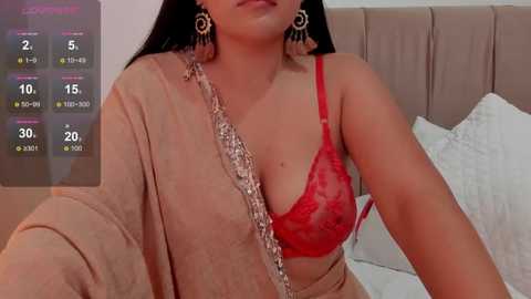 Media: Video of a woman with medium brown skin and long dark hair, wearing a red lace bra and gold earrings, lying on a beige bed, with a smartphone displaying a health app in the background.