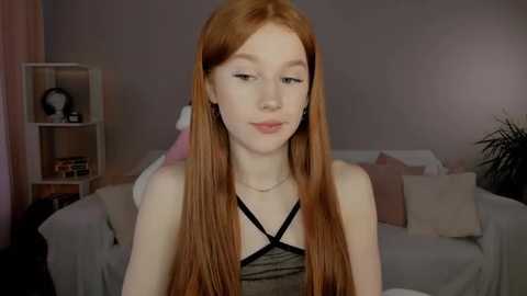 Media: Video of a young Caucasian woman with long, straight red hair, fair skin, and a slender physique. She wears a black camisole with thin straps. The background features a white couch with pillows, a bookshelf, and a plant.