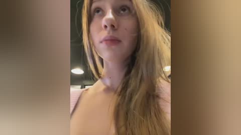 Media: Video of a young Caucasian woman with long, light brown hair, fair skin, and a slender build. She has a neutral expression and wears a pink top. The background is blurred, suggesting an indoor setting with soft lighting.