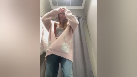 Media: Video of a young woman in a fitting room, adjusting a pink oversized sweater, wearing a gray tank top and blue jeans, with a white purse.