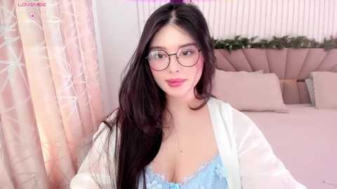 Media: Video of a young Asian woman with long black hair, wearing glasses and a light blue lace bra, standing in a pink bedroom.