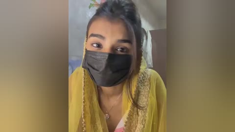 Media: Video of a young South Asian woman with medium skin tone, wearing a yellow saree with intricate golden embroidery, black face mask, and brown hair tied up. She gazes directly at the camera in a dimly lit room.