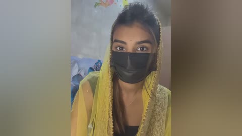 Media: Video of a young South Asian woman with medium skin tone, wearing a yellow and beige embroidered dupatta, black face mask, and black top, in a softly blurred background with floral patterns.