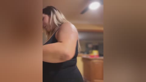 Media: A blurry video of a plus-sized woman with long blonde hair, wearing a black dress, seen from the side in a dimly lit, indistinct kitchen setting.