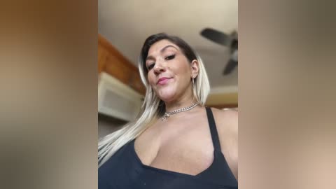 Media: A video of a Latina woman with long, straight black hair and blonde highlights, wearing a black halter top, standing in a kitchen with wooden cabinets and a ceiling fan.