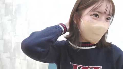 Media: Video of an East Asian woman with straight, shoulder-length brown hair, wearing a navy blue sweater with red and white accents, a beige mask, and a white headband, adjusting her hair against a softly lit, white background.