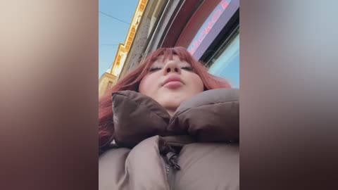 Media: Video of a young woman with red hair and light skin, wearing a puffy brown jacket, taken from a low angle, showing her chin and mouth. Background includes a building with red and blue signage.