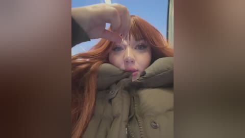 Media: Video of a young woman with long, straight red hair, wearing a green jacket, sticking her tongue out playfully while indoors with a clear blue sky visible through a window.