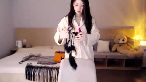 Media: Video of a young East Asian woman with long black hair in a white bathrobe, trimming a black dog's tail on a white bed in a modern bedroom with a beige headboard and plush teddy bear.