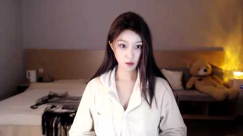 Media: A video of an Asian woman with long, straight black hair, fair skin, and light makeup, wearing a white button-up shirt, standing in a dimly-lit bedroom with a beige bed, plush teddy bear, and a glowing nightlight.