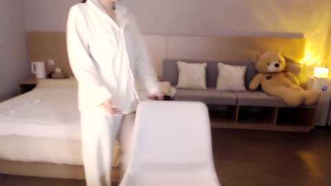 Video of a person in a white bathrobe, standing near a bed with white linens, a beige headboard, and plush toys.