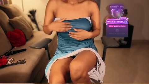 Media: Video of a woman in a strapless blue dress, revealing her large breasts, sitting on a beige couch in a dimly lit living room. A purple virtual reality headset with a game display is in the background.