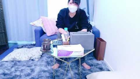 Media: Video of an Asian woman with a mask, dark hair, wearing a black sweatshirt, seated at a glass table with a grey suitcase, documents, and a brown bag. Blue chair, fluffy rug, and white curtains in the background.