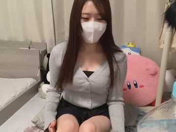 Media: Video of an East Asian woman with long brown hair, wearing a white mask, gray cardigan, and black shorts, seated on a bed in a bedroom with a plush Kirby plushie and a fan.